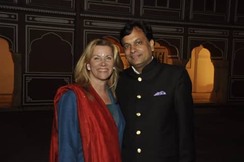 Two-shot: Gayle in red shawl with man in black bandhgala, palace backdrop